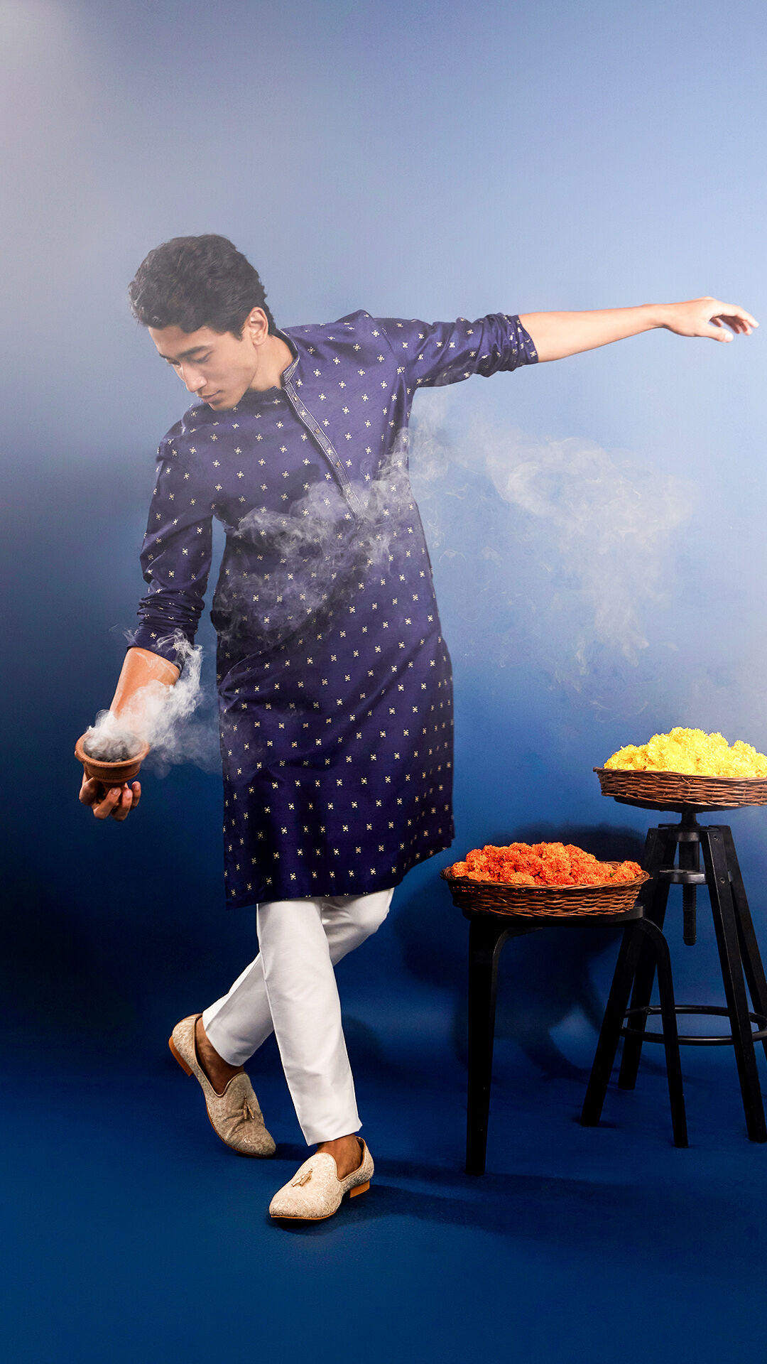 Man performing festive ritual in navy blue kurta with golden motifs, paired with white pants and ethnic footwear, surrounded by marigold flower baskets